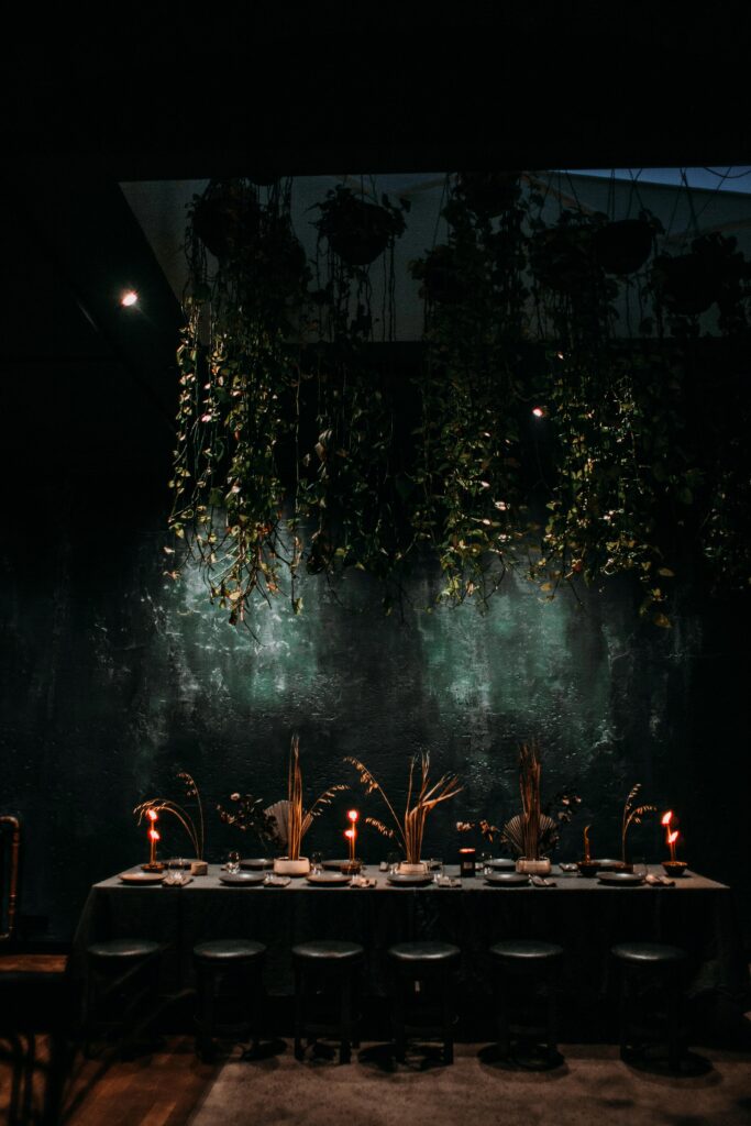Interior of contemporary dark restaurant with long set dining table with burning candles under hanging verdant plants